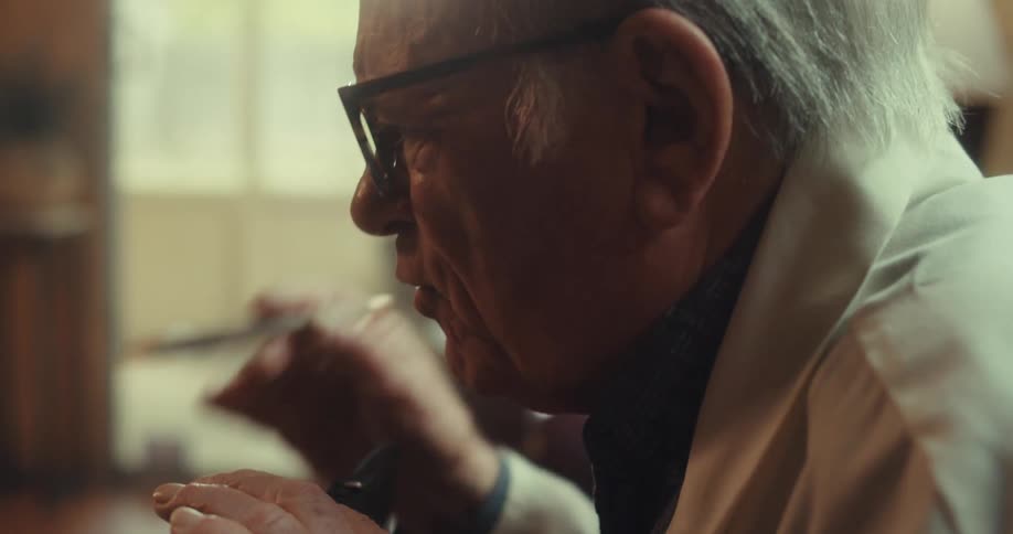 Old man with gray hair and black framed glasses is speaking to someone a bit aggressive he is gesturing as he talks with his hand which holds a pen the man is in a naturally lit possible living room with a white bricked fire place and wooden floors