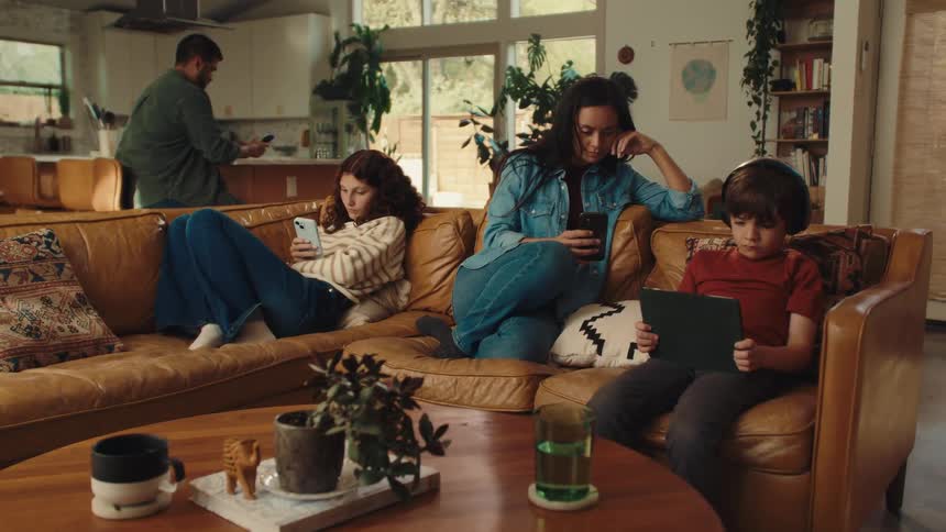A woman, a teenager, and a child sitting on the couch focused only on their phones and tablet, and in the background a man is sitting in the kitchen looking at his phone.