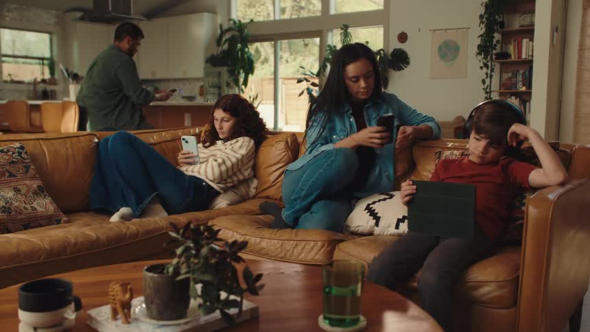 A family sits quietly together on a living room couch each person absorbed in their own digital device while a man prepares something in the kitchen behind them