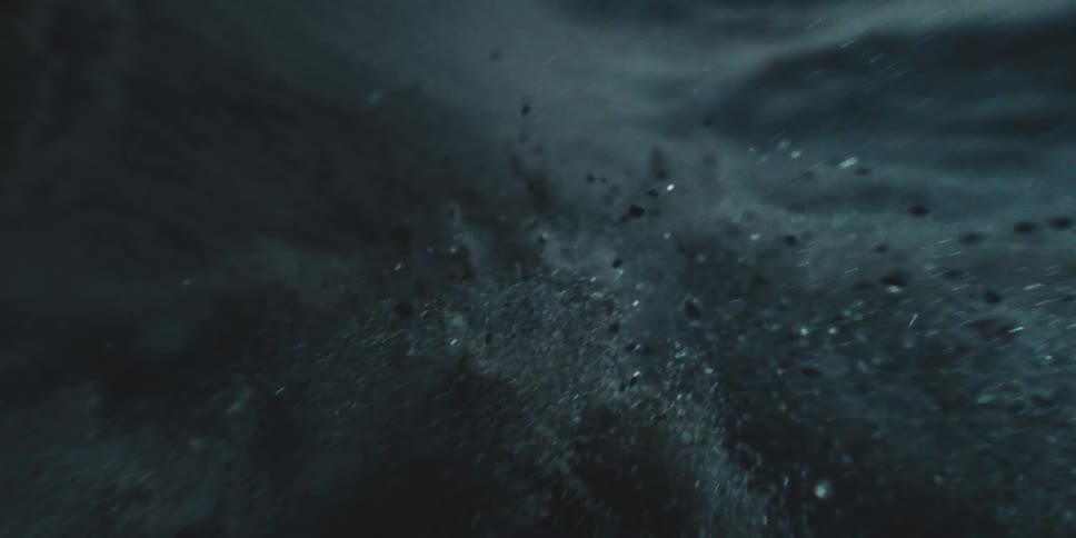 Dark rock particles swirl in liquid or air creating a moody abstract scene as the camera slowly moves forward through a cloudlike burst
