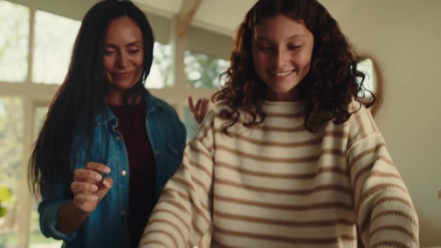 Mother and daughter smile while the older woman guides her daughter in their kitchen