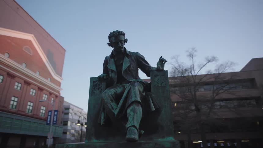 A weathered bronze statue of a man sits alone in an urban setting under a muted twilight sky