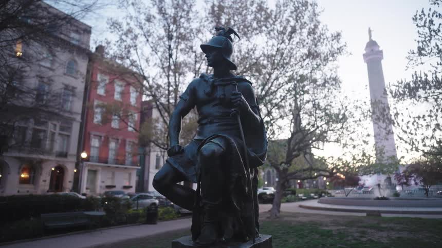 A stone sculpture of a person wearing a hat sitting on a stone block in a square