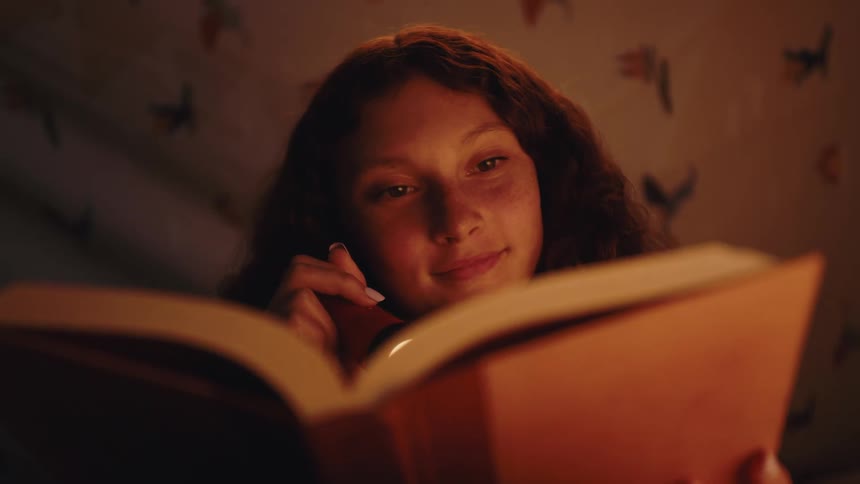 A girl reading a book while holding a flashlight to see the letters