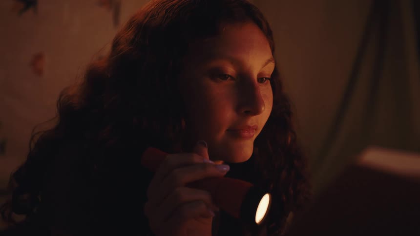 The teenager is using a flashlight to read a book in the dark room