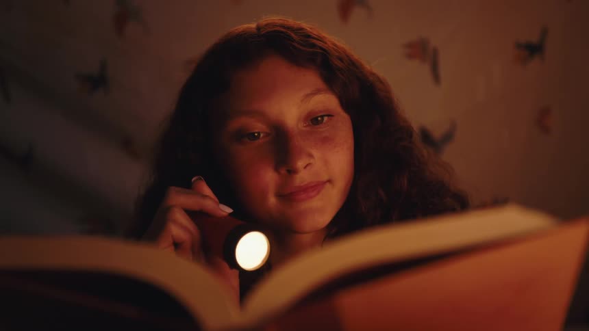 Teenage girl reading a book holding a flashlight in dark room