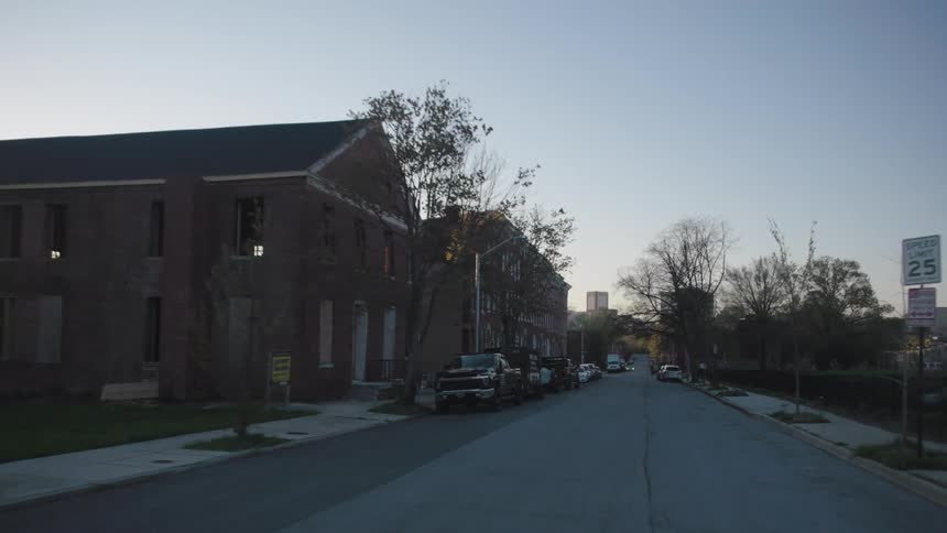 A dimly lit quiet street scene at dusk with several large buildings lining the road under a clear pale blue sky