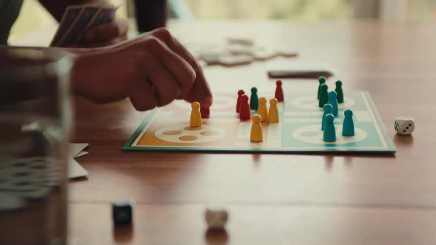 A person is playing a board game with colorful pieces on a wooden table with dice and cards also visible