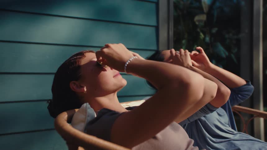 Two women smiling while sitting in chairs on the porch and placing cucumber slices on their eyes