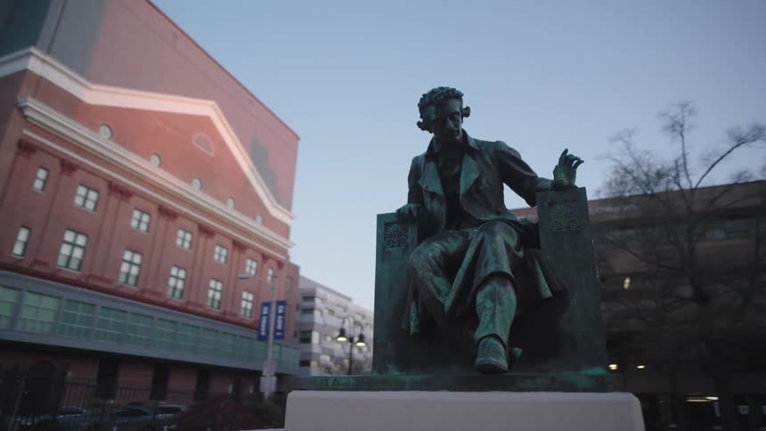 A footage of a mans statue sitting on a chair just in front of the brown buildings