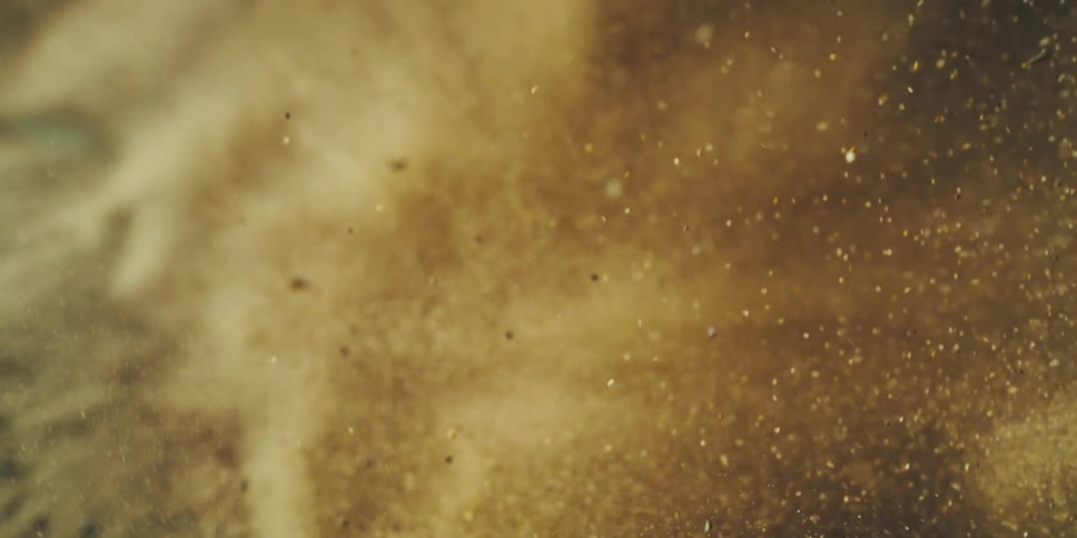 A dramatic closeup captures particles of golden powder swirling and dispersing through the air in slow motion