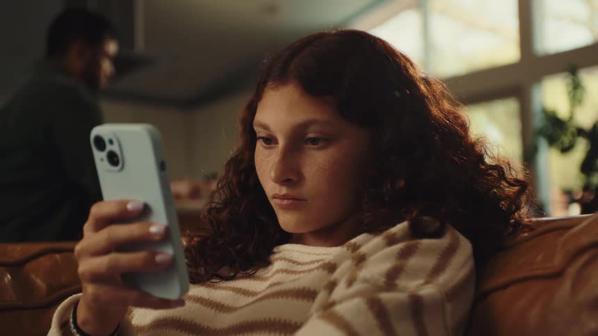 A teenager / young lady sitting on a couch indoor holds a smartphone at daytime