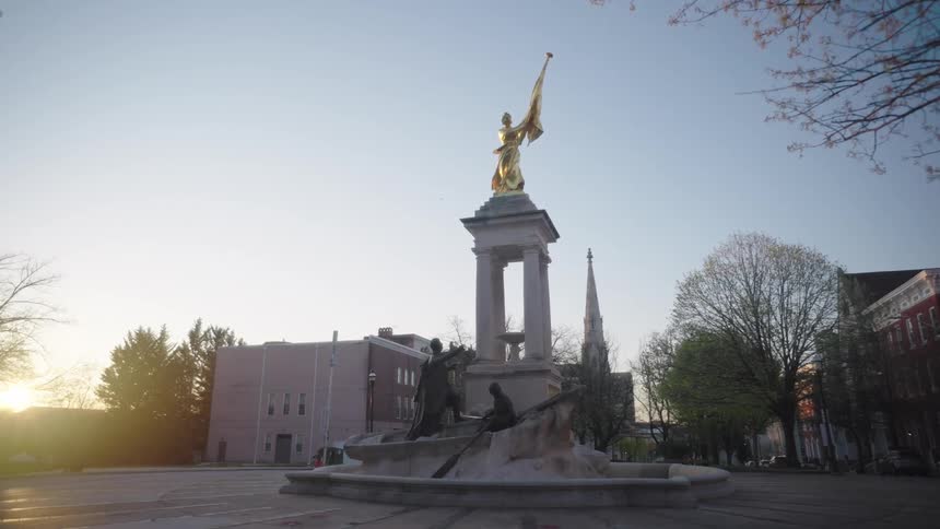 Monument with a golden statue of a woman holding a flag on top of a column surrounded by sculptures and urban buildings in the background