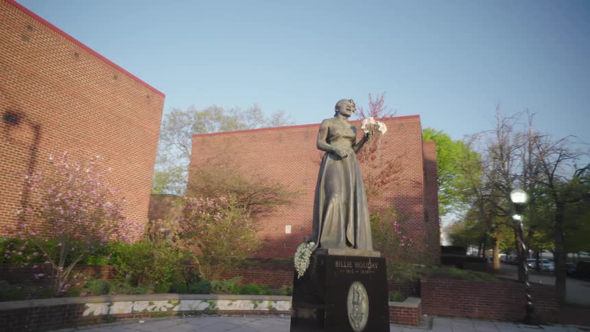Statue standing in a plaza near a street holds a bouquet of fresh flowers