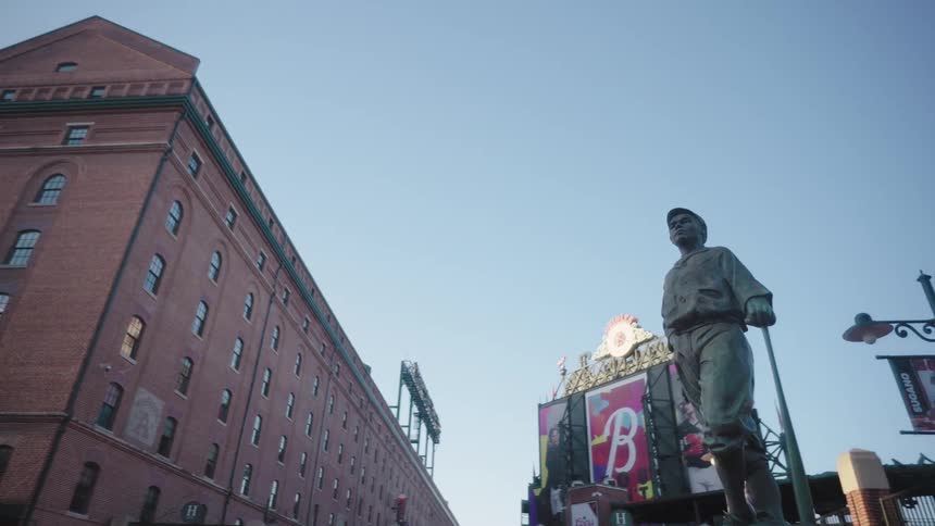 A towering statue of a baseball player stands in front of a historic red brick building under a clear blue sky in a cityscape setting