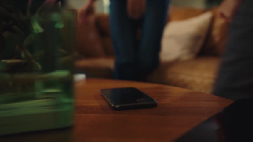 A smart phone on a table and three young people on a couch later they leave and a girl places another smart phone on the table