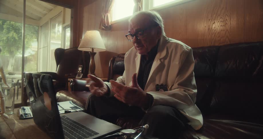 Man in white coat provides video conference service sitting on sofa with a stethoscope on the table