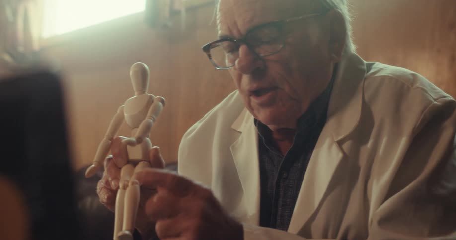 An elderly man in a lab coat explains or demonstrates something using a wooden mannequin likely in a thoughtful and educational context
