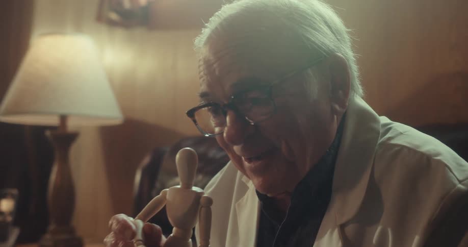 A man wearing glasses and a long coat sitting on a couch in a living room talks at a laptop in front of him while manipulating a wooden articulated doll