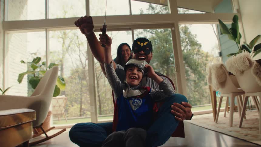 A child in a superhero costume leads a playful indoor ride cheered on by joyful parents
