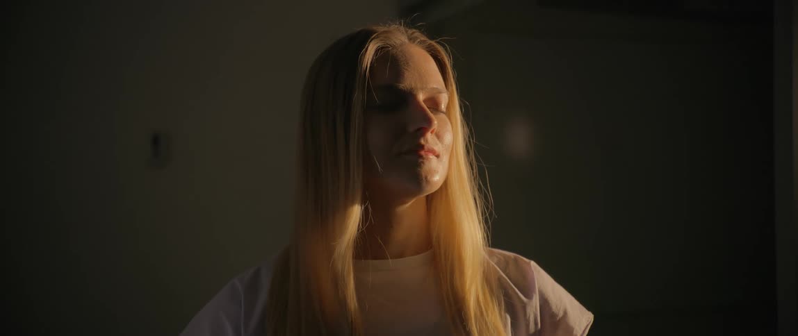 A young blonde female with long hair stands with her eyes closed as light reflects softly on her face