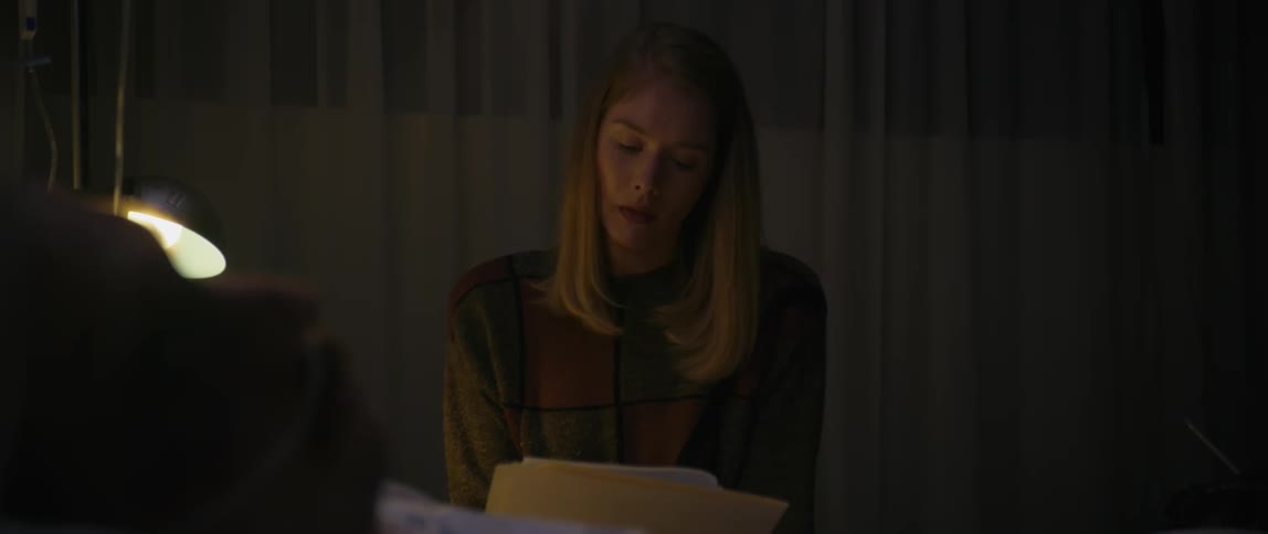 A young blonde woman is sitting in a partially dark indoor environment reading a paper