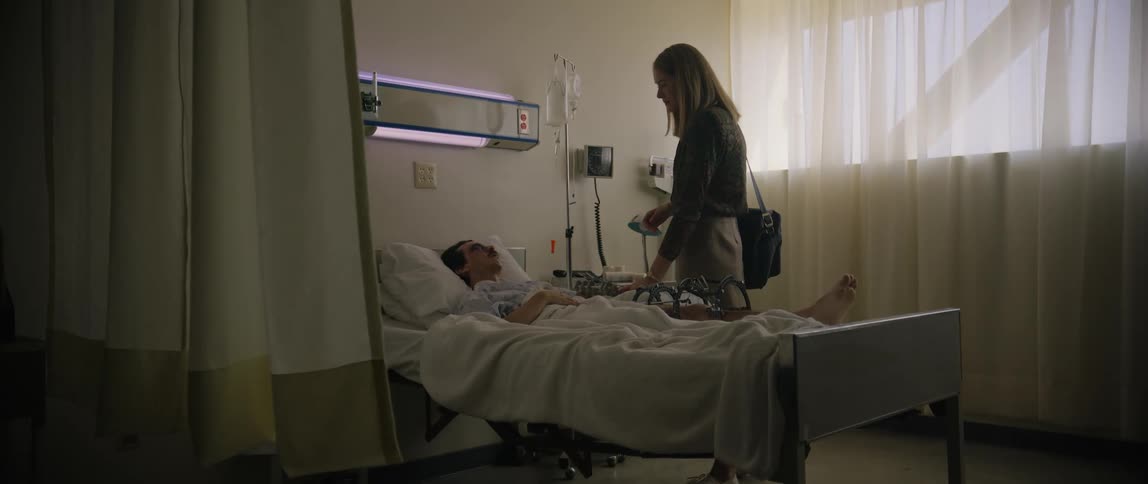 A woman visiting a bedridden man in a hospital bed with a drip bottle by his side and daylight streaming through the window