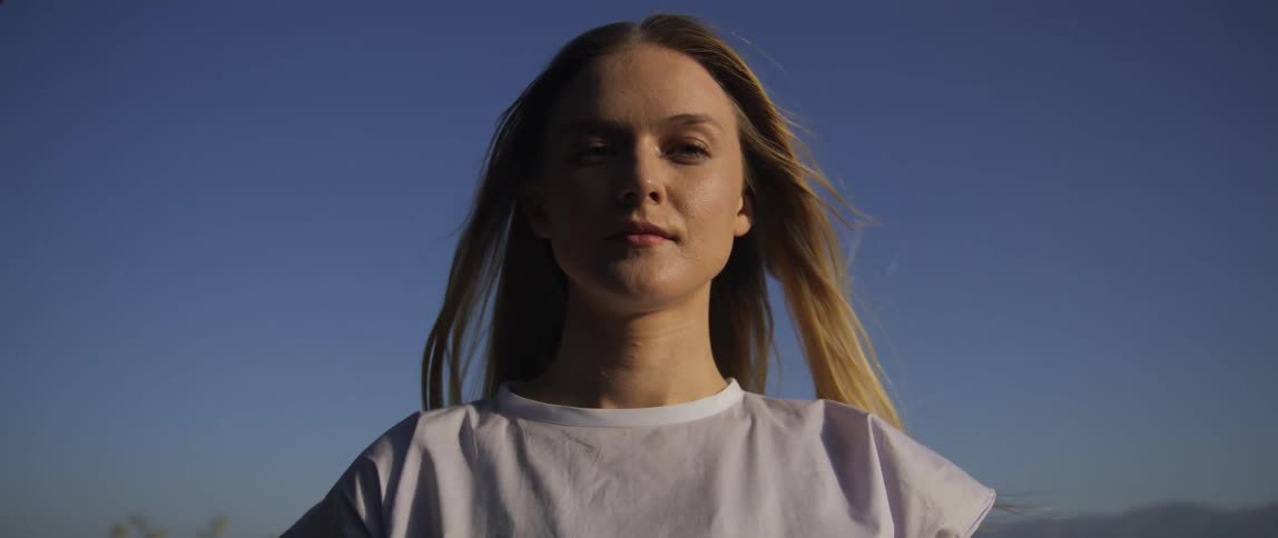 A young woman stands calmly outdoors with her eyes closed and then opens them appearing peaceful and contemplative under a clear blue sky