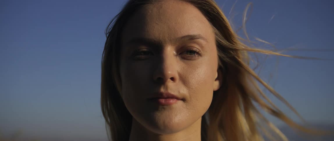 A young woman stands still and gazes directly into the camera with a calm neutral expression under natural light with the wind blowing her hair against a clear blue sky