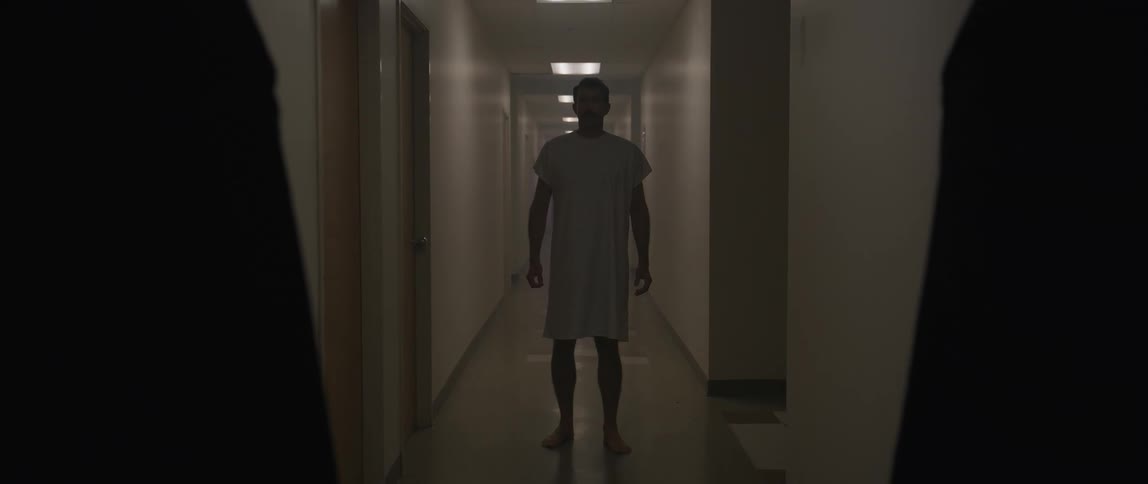 A man in hospital scrubs walks out of a room into a hallway and then stands there watching two people in front of him