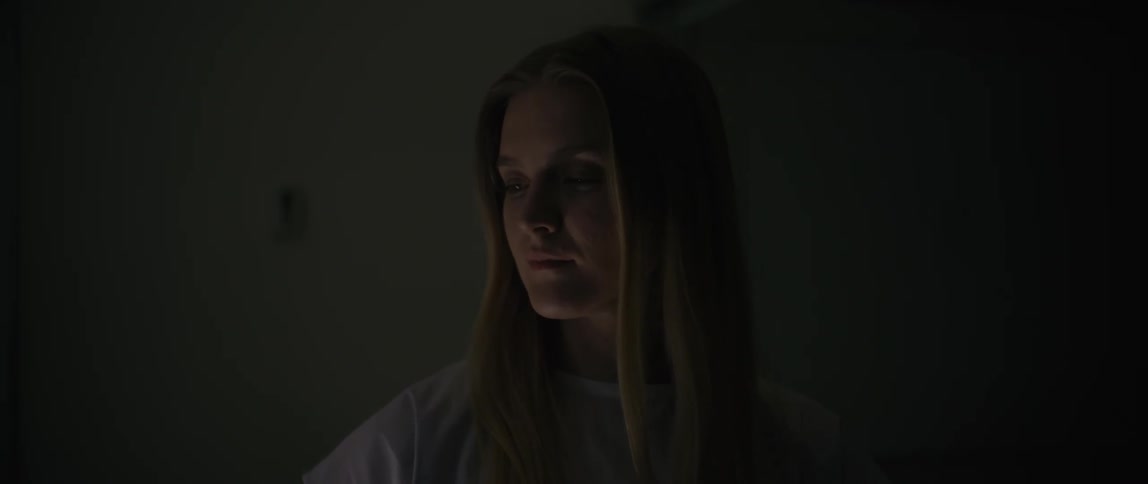 A young woman with long blonde hair is softly lit in a dimly lit interior gazing thoughtfully offcamera