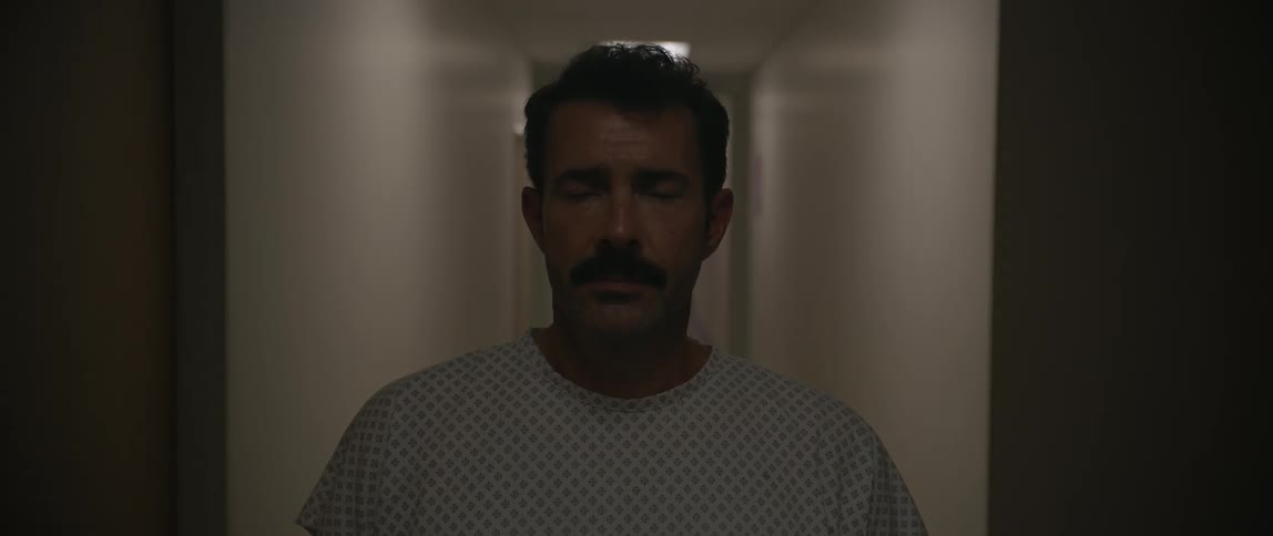 A man stands in the middle of a dark corridor that gets illuminated as he blinks and looks from side to side