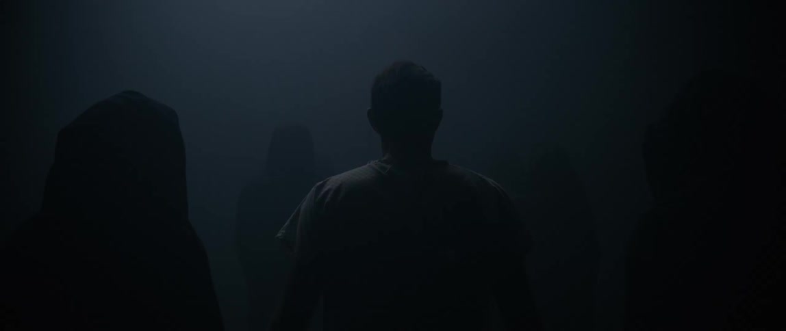 A man is nudged from behind by a dark figure as he walks away along a dark corridor