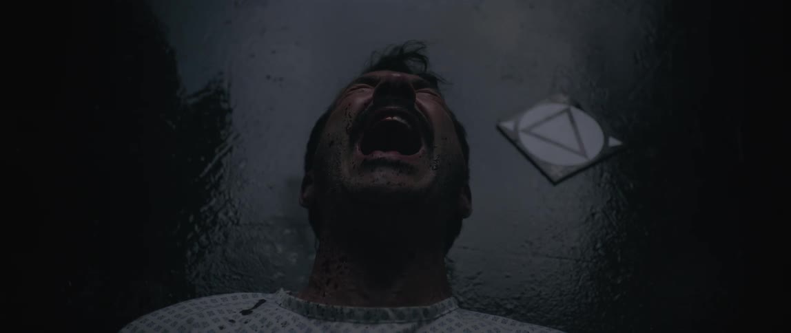 A distressed man lies on the floor in a dark eerie setting seemingly in pain or anguish viewed from directly above