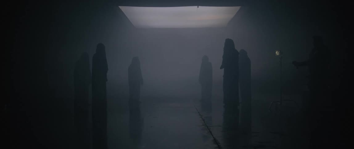 Hooded people stand in a circle under a skylight in a dark foggy environment