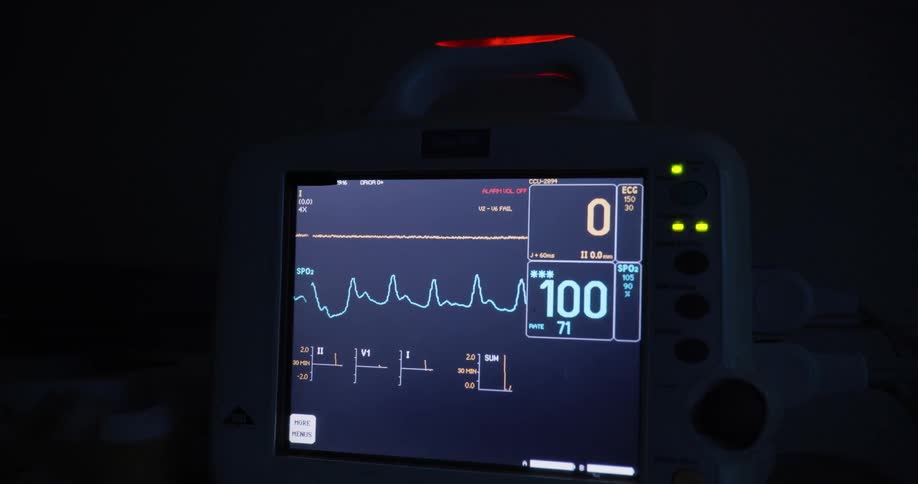 A dimly lit room shows a closeup of a medical monitor displaying heart rate and oxygen levels