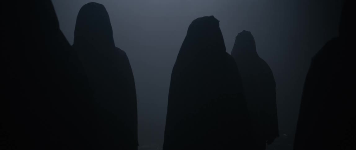 A group of people dressed in black clothing walk along a dimly lit corridor at night