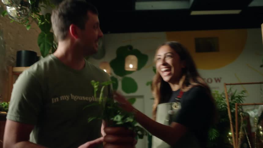 A smiling woman hands a man a potted plant who then walks away as the woman picks up a new empty pot off the desk
