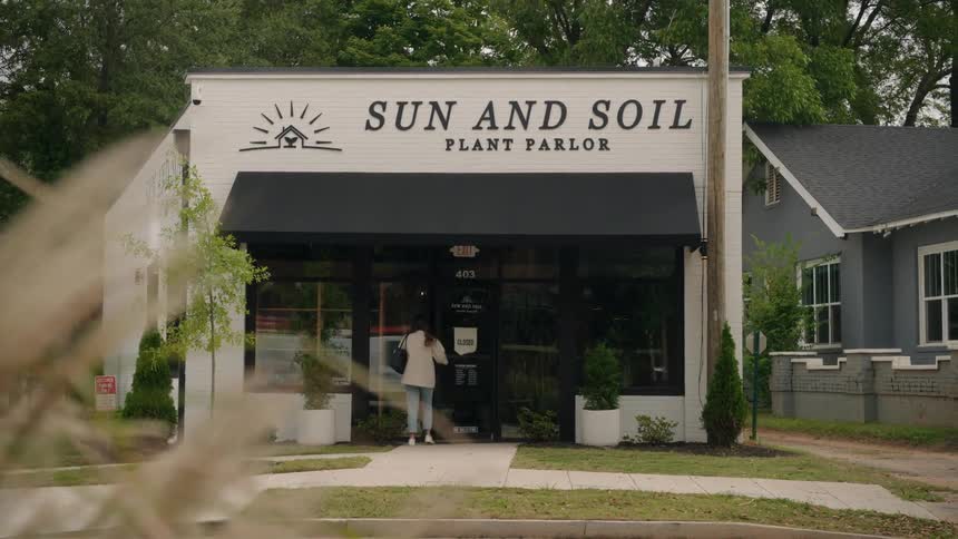Woman opens the door to a plant store near a busy street and flips the welcome sign to open