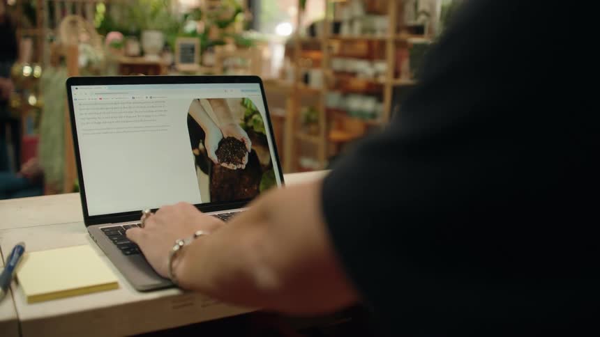 A person is browsing a plantthemed website on a laptop in what appears to be a cozy plantfilled store or workspace