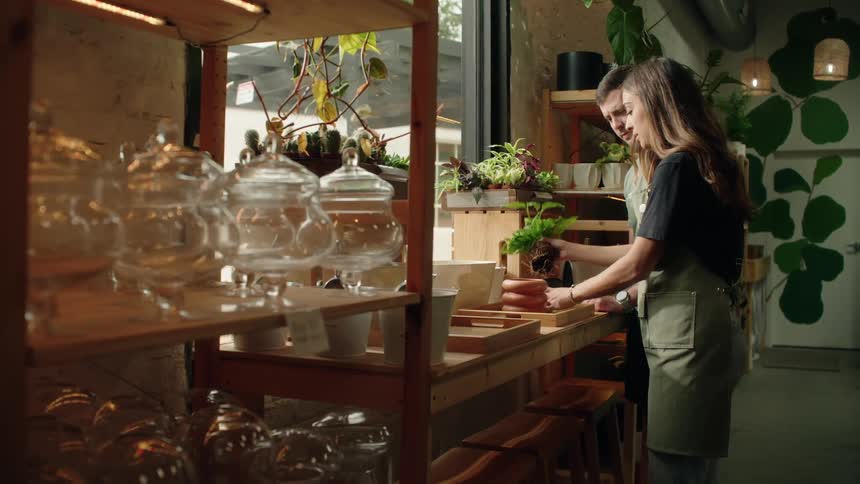 A man and woman are working together to pot plants in a cozy indoor gardening shop filled with glass containers and greenery