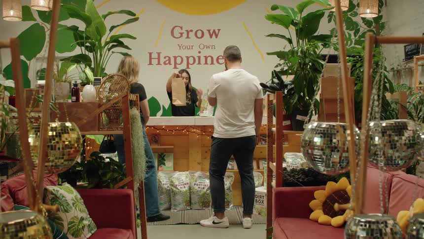 A young woman places a flower in a bag and hands it to a blonde woman on the other side of a counter before handing her change while a man wearing white stands nearby