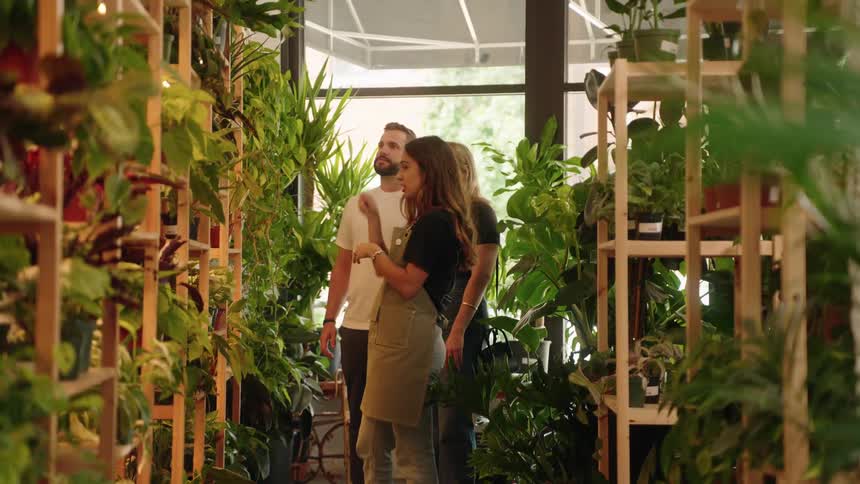 Two customers are being assisted by a female employee amidst a vibrant display of plants and foliage in a greenhouse like setting