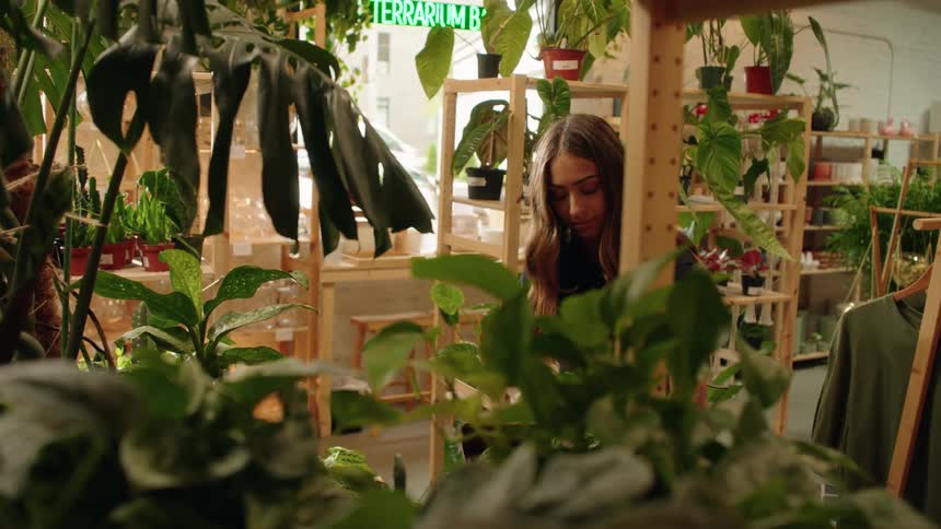 A young woman is tending to plants in an indoor plant shop filled with lush greenery and wooden shelving