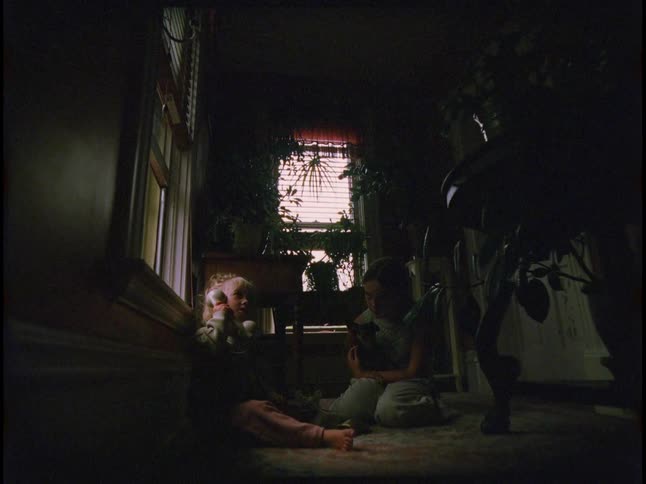 A child and toddler share a quiet moment on a rug in a dim plantfilled room lit by window light