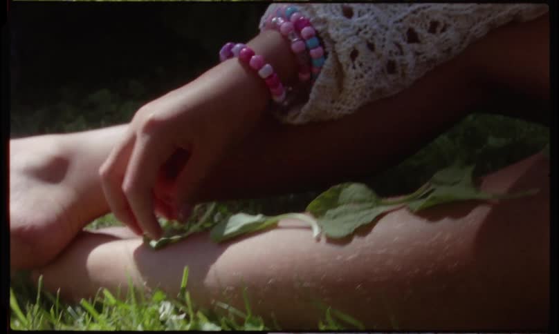 A young girl plays in a sunlit garden gently placing leaves on her leg in a calm and contemplative moment