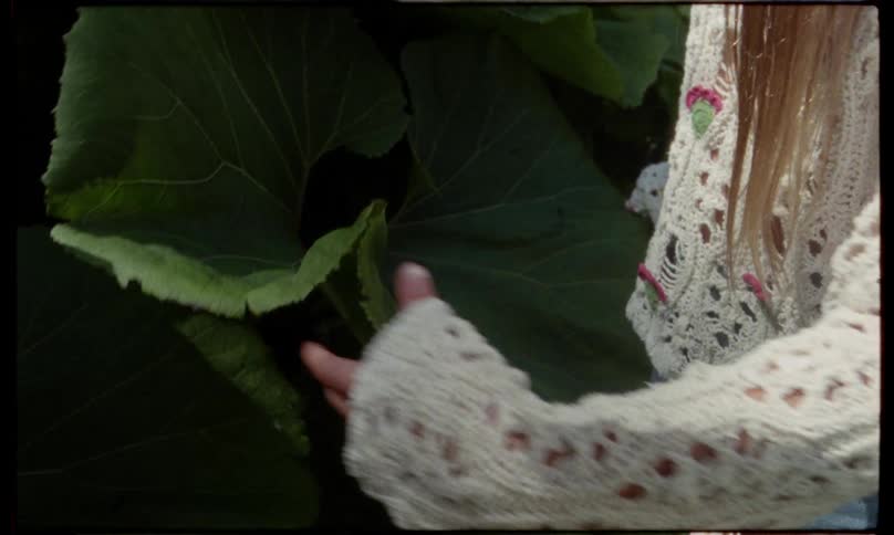 A young woman gently touches large green leaves in a garden while wearing a crocheted sweater with soft sunlight casting natural tones