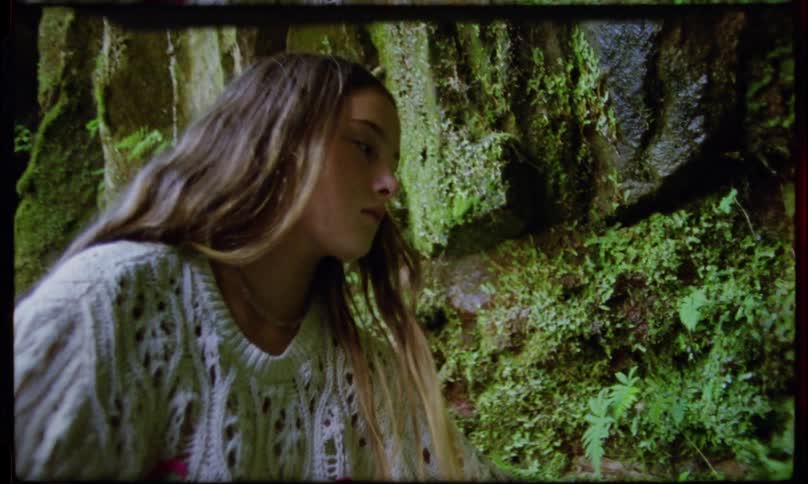 A teenage girl in a white crocheted sweater explores a lush mosscovered outdoor setting reaching out to touch the natural rock and foliage around her