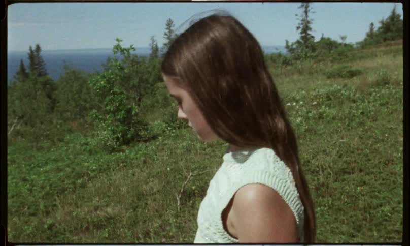Young woman walks down a grassy slope near a coast dotted with groves of trees on a summer day