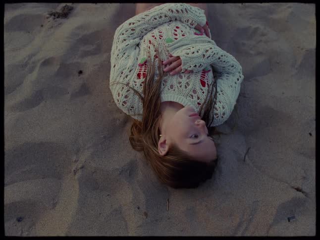 Young girl sitting up facing the shoreline then lying down on the sand